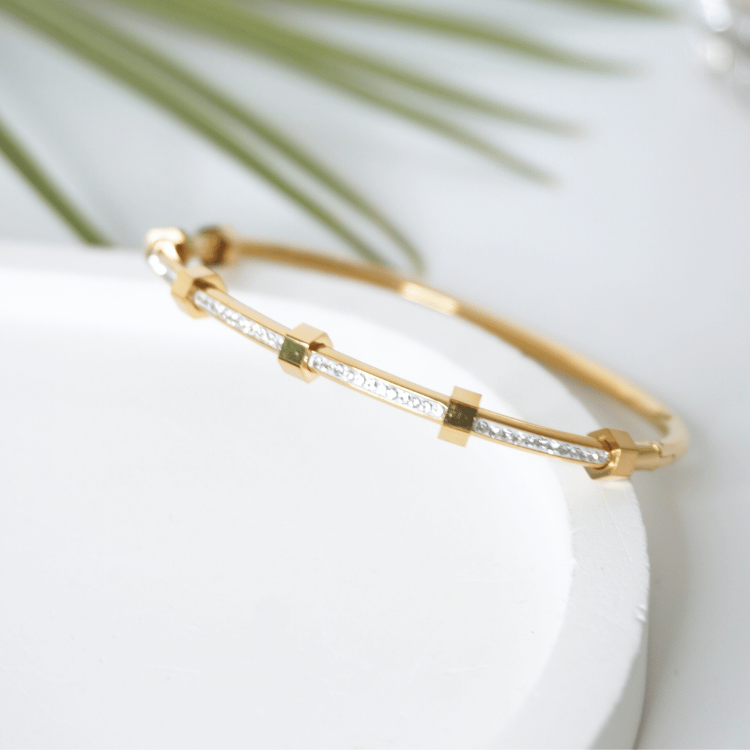 Gold bracelet with embedded stones on a white surface with green leaves in the background